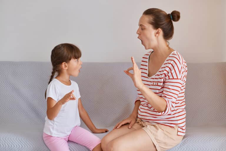 Side view of woman teacher wearing striped shirt and shorts sitting on sofa with little girl, preschooler kid practicing correct pronunciation with a female speech therapist.